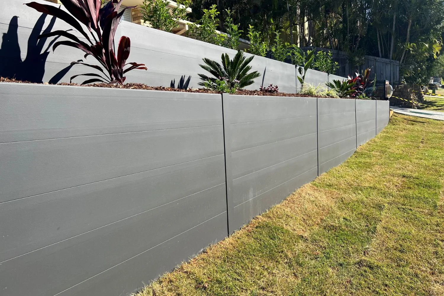 A grey concrete sleeper retaining wall with tropical planting alongside a paved area