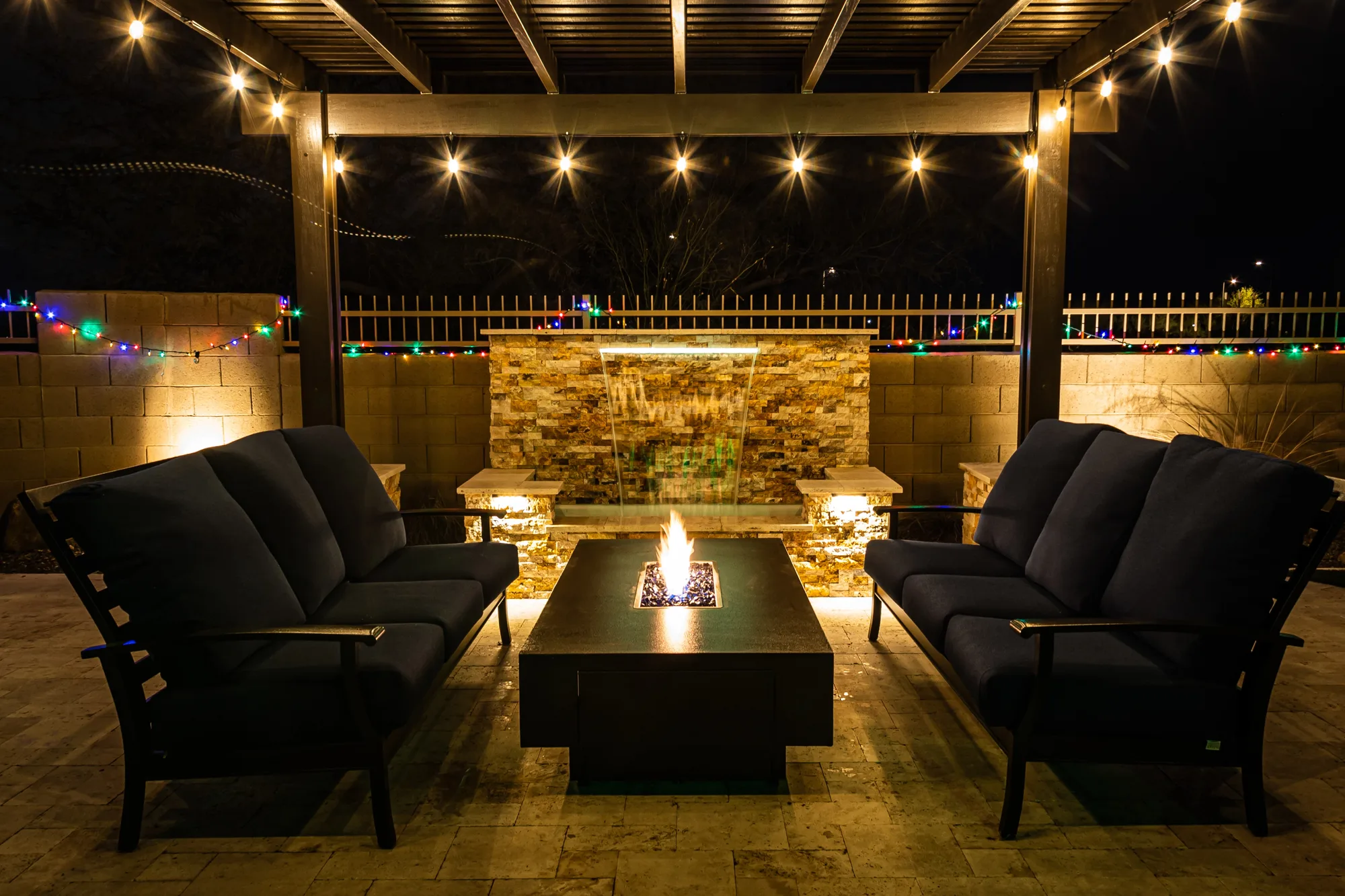 A stone fire pit lounge area with a water feature and string lighting